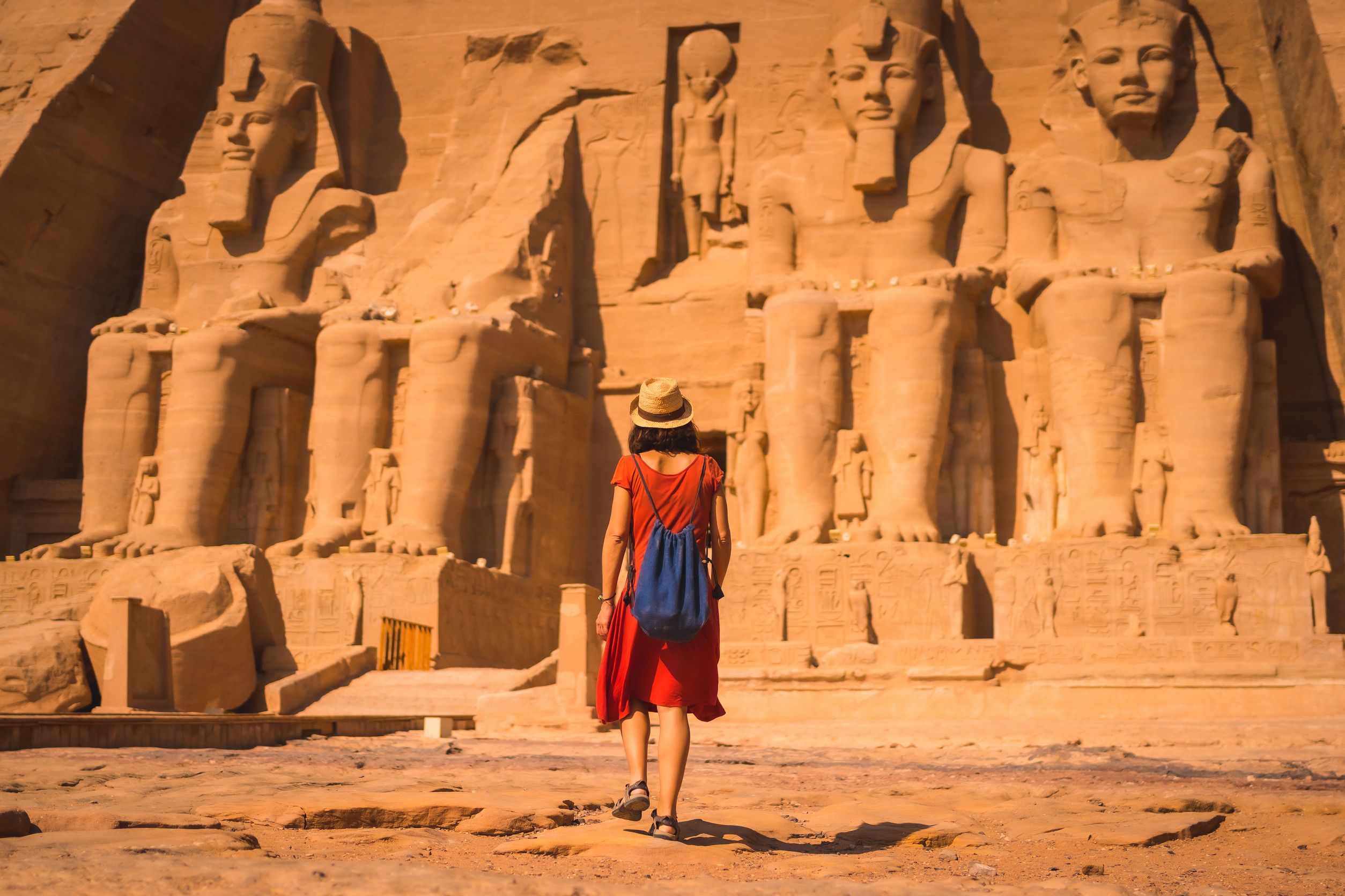 Young Tourist In A Red Dress And Straw Hat Walking Towards The Abu Simbel Temple In Southern Egypt